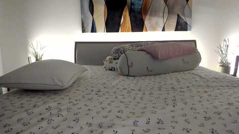 Media: Video of a minimalist, modern bedroom with a large bed featuring a white, patterned duvet, two white pillows, and a rolled-up pink blanket. The headboard is upholstered in light gray fabric, and behind it hangs a colorful abstract painting. The room has two bedside tables with small potted plants, each illuminated by a soft light.