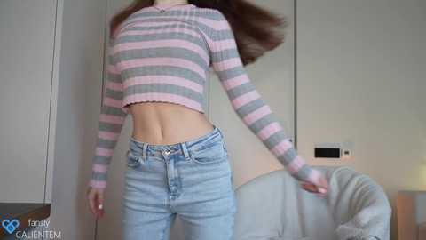 Media: Video of a young woman with long brown hair, wearing a pink and gray striped crop top and light blue jeans, dancing in a modern, minimalistic room with white walls and a couch.