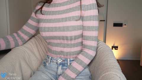 Media: Video of a woman with medium breasts, wearing a pink and gray striped sweater and light blue jeans, sitting in a beige armchair in a modern, dimly lit room with a door and light switch.