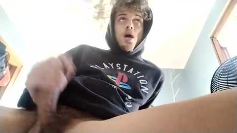 Media: A young man with light skin, short brown hair, and a PlayStation hoodie, is seen from below, holding his erect penis. He is in a bedroom with a fan and wooden furniture.