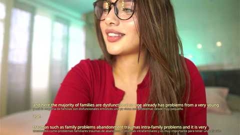 Media: Video of a smiling woman with long brown hair, wearing black-rimmed glasses, red cardigan, and makeup. Text overlay discusses family problems. Background features a softly lit bedroom with large windows.