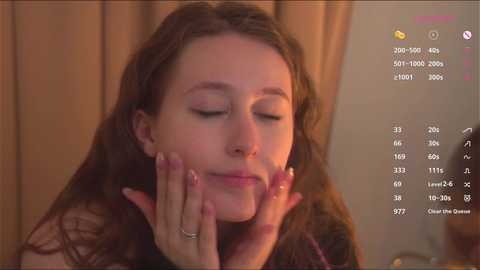 Media: Video of a young woman with long brown hair, light skin, and closed eyes, gently touching her face. Background features beige curtains and a blurred wooden surface.