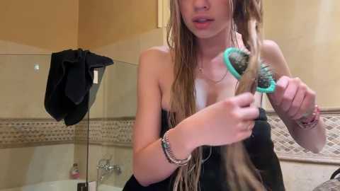 Media: Video of a young woman with long, wet, blonde dreadlocks, holding a teal hairdryer in a beige-tiled bathroom with a black towel on a glass shelf.