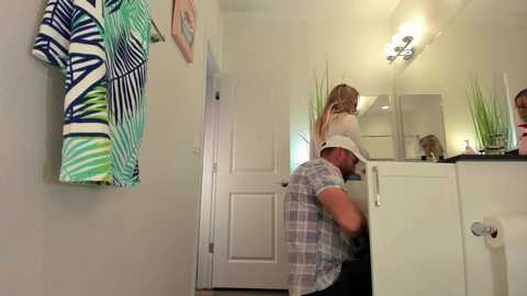 Media: Video of a woman with long blonde hair in a white shirt, standing in a white-tiled bathroom with a mirror, green plants, and colorful clothes hanging on the wall.