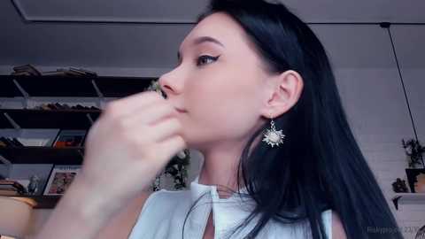 Media: Video of a fair-skinned woman with long black hair, wearing a white blouse, and sunflower earrings, in a modern, cluttered room with shelves and potted plants.