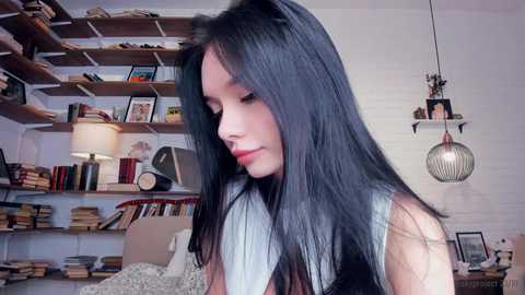 Media: Video of a young woman with long black hair, wearing a black tank top, standing in a cozy, book-filled room with wooden shelves, a lamp, and a hanging light fixture.