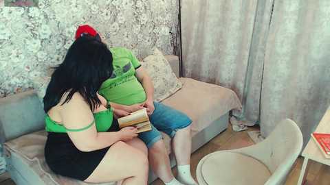 Media: A video of a curvy woman with black hair, wearing a green top and black shorts, reading a book while seated on a beige sofa. Her partner, in a green t-shirt and blue shorts, sits beside her. The room has a floral-patterned wallpaper and beige curtains.