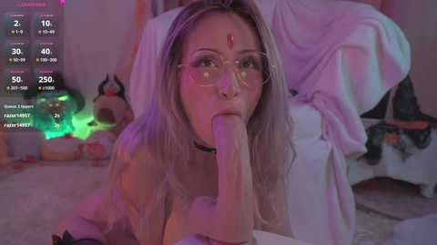 Media: A video of a blonde woman with glasses, wearing a choker, performing oral sex on a large, erect penis, set in a dimly lit, cluttered room with a green light and stuffed animals.