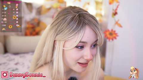 Media: A video of a young, fair-skinned woman with long, blonde hair and light makeup, smiling softly. Background features a cozy, colorful room with floral decorations and a bed. Watermark \"SunnySonic2\" visible.