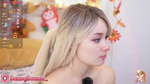 Media: Video of a young, light-skinned woman with blonde hair, wearing makeup, looking contemplative in a bedroom with autumn decorations.