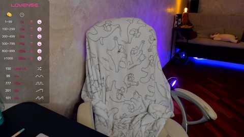 Media: Video of a woman wearing a white blanket with cartoon character sketches on a gaming chair in a dimly-lit room with a wooden floor, a table, and a TV screen displaying \"LIVEVIEW\" in the top left corner.