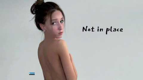 Media: Video of a young, fair-skinned woman with brown hair tied in a bun, topless, looking over her shoulder, with \"Not in place\" text in black.