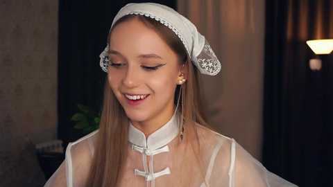 Media: Video of a young woman with fair skin, long blonde hair, and delicate features wearing a white lace headpiece and sheer veil, smiling softly. Background shows a dimly lit indoor setting with a soft light source.