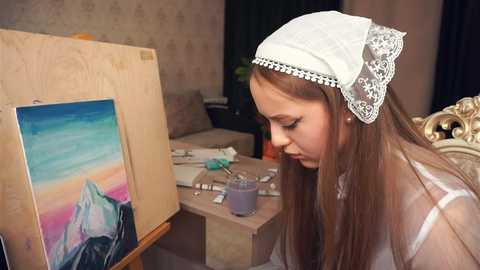 Media: Video of a young woman with long brown hair, wearing a white lace headscarf, painting a vibrant seascape on a wooden easel in a cozy, dimly-lit living room.