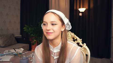 Media: Video of a fair-skinned young woman with long brown hair, wearing a white lace veil and delicate dress, sitting at an ornate wooden table in a softly lit, cozy room with dark curtains and a potted plant.