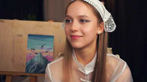 Media: Video of a young Caucasian woman with straight, light brown hair, wearing a white lace headband and a sheer white blouse. She smiles softly. Background includes an easel with a colorful landscape painting and a dark curtain.