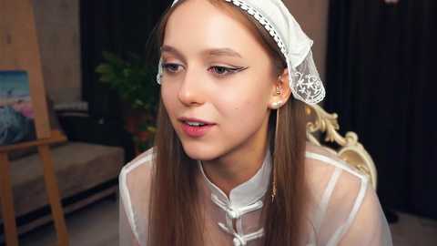 Media: Video of a young Caucasian woman with light skin, brown hair, and a white lace veil. She wears a white, semi-transparent dress with gold buttons. The background shows a painting of a beach and a green plant.