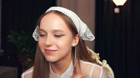 Media: Video of a fair-skinned young woman with long brown hair, wearing a white lace headscarf and a sheer white blouse, smiling gently, in a softly lit indoor setting.