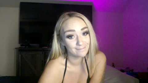 Media: A video of a smiling young woman with blonde hair, wearing a black top, sitting in a dimly-lit room with a TV and purple lighting.