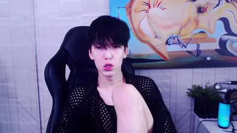 Media: Video of a pale, short-haired individual with a pierced nose and bright pink lipstick, wearing a black mesh top, sitting in a black gaming chair. Artistic background with abstract floral painting and gaming accessories.