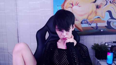 Media: Video of a young, pale-skinned, androgynous person with short black hair, wearing a black mesh top, sitting in a black gaming chair. Background features a colorful abstract painting and a potted plant.