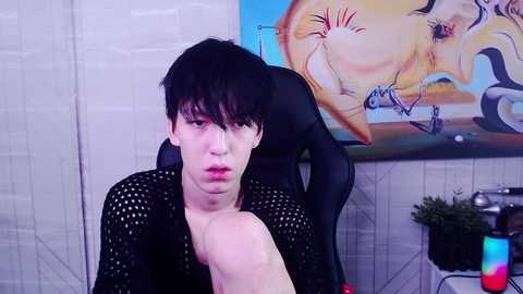 Media: Video of a young, pale-skinned, androgynous person with black hair, wearing a black mesh shirt, sitting in a black gaming chair, in a room with a colorful abstract painting and gaming equipment.