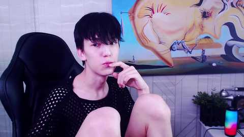 Media: Video of a slender, pale-skinned young man with short black hair, wearing a black mesh shirt, sitting pensively in a black gaming chair. Background features a colorful, abstract painting and a green plant.