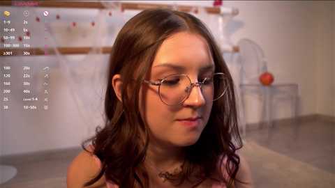 Media: Video of a young woman with long brown hair and glasses, wearing a pink top, in a home setting with a calendar and basketball hoop in the background.