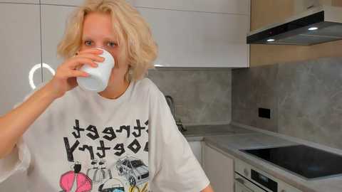 Media: Video of a blonde woman with wavy hair, wearing a white t-shirt with graphic design, drinking from a white mug in a modern kitchen with white cabinets, black countertop, and stainless steel appliances.