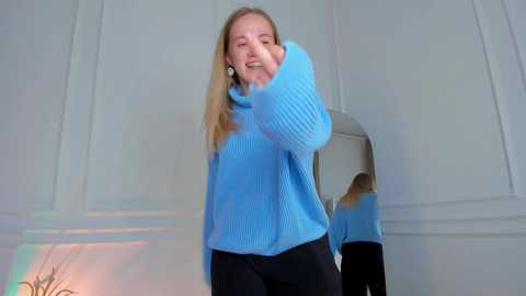 Media: A video of a smiling blonde woman with shoulder-length hair, wearing a blue sweater and black pants, standing in a white room with a reflective surface.