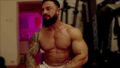 Media: Video of a shirtless, muscular man with a full beard and tattoos on his right arm, wearing white shorts, standing in a dimly lit gym with equipment in the background.