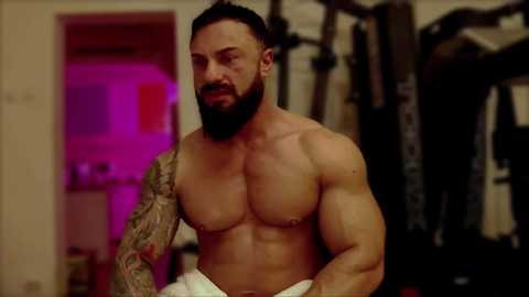 Media: Video of a muscular, tattooed, bearded man with a white towel around his waist, standing in a dimly lit gym.