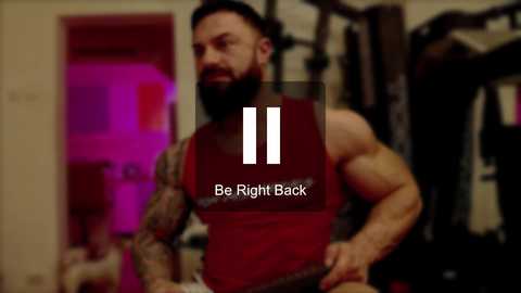 Media: Video of a muscular, tattooed man with a beard working out in a gym, wearing a red tank top. Text overlay: \"Be Right Back.\