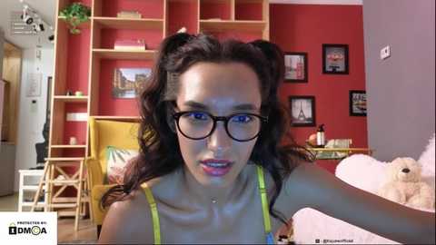 Media: Video of a young woman with dark hair styled in pigtails, wearing black-rimmed glasses and a yellow spaghetti-strap top, sitting in a cozy room with red walls, wooden shelves, and a plush teddy bear.