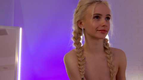 Media: Video of a young, fair-skinned blonde woman with twin braids, wearing a black top, smiling softly. Background features a purple-lit wall and a modern, minimalist interior.