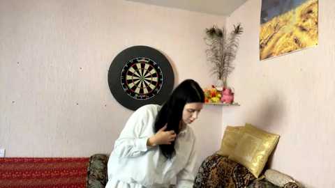 Media: A video of a young woman with long black hair, wearing a white shirt, adjusting her hair while seated on a patterned couch in a room with a dartboard and floral artwork.