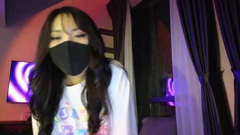 Media: Video of an Asian woman with long black hair, wearing a black face mask, white T-shirt with colorful graphic, standing indoors with dark curtains and purple lights in the background.