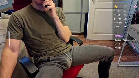 Media: Video of a fit, fair-skinned man with short hair, wearing a green t-shirt and gray pants, sitting in a red chair. Background includes a white door, wooden floor, and a computer screen showing a custom avatar editing tool.