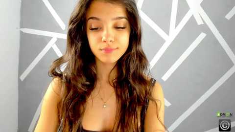 Media: A video of a young woman with long, wavy brown hair, wearing a black top, standing in front of a gray wall with white geometric patterns. She has a neutral expression.