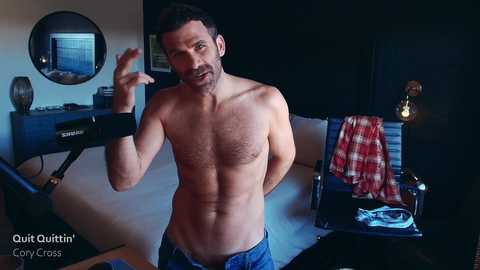 Media: Video of a shirtless, muscular white man with short dark hair and a beard, wearing blue jeans, gesturing in a dimly-lit, modern living room with a black leather armchair, a plaid blanket, and a dark wooden desk.