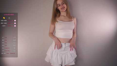 Media: A video of a smiling, fair-skinned young woman with long blonde hair, wearing a white crop top and ruffled skirt. She stands against a plain gray background, with a digital overlay showing social media metrics.