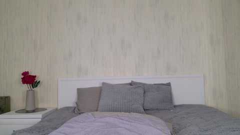 Media: A video of a modern, minimalist bedroom with a white headboard, gray bedding, and a vase with red flowers on a white nightstand.