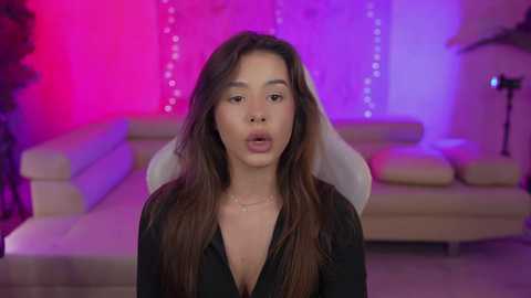 Media: A video of a young woman with long, brown hair, wearing a black shirt, in a modern living room with purple and pink LED lights.