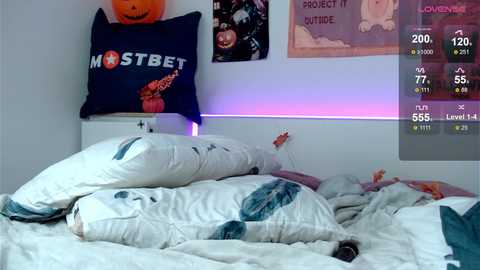 Media: Video of a messy bedroom with a white bed covered in blankets, a dark pillow with \"MST3K\" text, a Halloween pumpkin on a shelf, and a purple light strip.