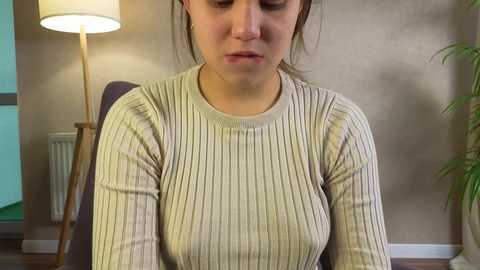 Media: Video of a young woman with fair skin and brown hair in a ponytail, wearing a beige ribbed sweater, seated indoors, with a lamp and potted plant in the background.