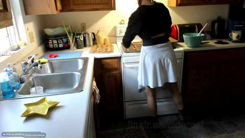 Media: Video of a woman in a white apron washing dishes in a small, cluttered kitchen with wooden cabinets and a sink filled with dishes.