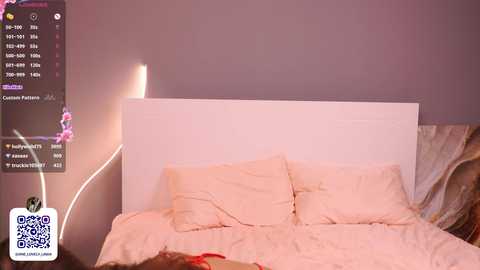 Media: Video of a minimalist bedroom with a white bed, beige pillows, and a dimly lit purple wall. A smartphone with a QR code and a pink floral wallpaper is displayed in the top left corner.