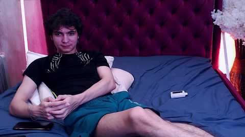 Media: Video of a young man with curly hair, wearing a black t-shirt and green shorts, lying on a bed with a dark blue sheet.