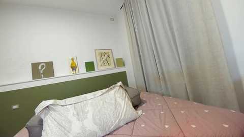 Media: Video of a minimalist bedroom with a green headboard, gray and white patterned pillows, and a pink bedspread with heart motifs. Artwork and sheer curtains adorn the walls and windows.