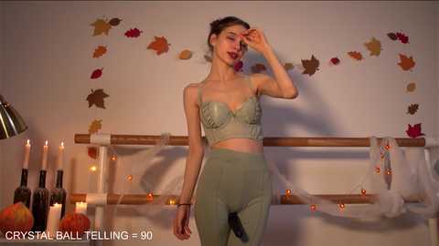 Media: Video of a slender, light-skinned woman in green lingerie and high-waisted pants, touching her face, in a dimly lit room with autumn-themed decorations and lit candles.